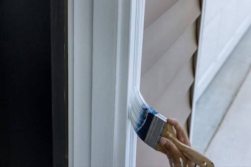 Close-up of wood trim around a door frame being painted white, with tan siding in view