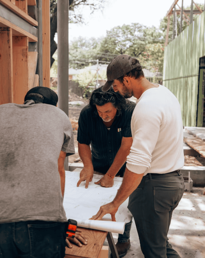 Three construction workers stand around a table with construction plans in the center.
